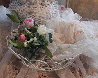 May include: A white, wirework basket holds a floral arrangement of pink and white tulips, green leaves, and small white flowers. The basket sits on a lace-covered surface, with a larger basket in the background.