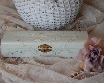 May include: A rectangular off-white box featuring raised floral relief patterns and a central gold-toned metal latch. The box has a distressed finish with visible wear and is accompanied by a single artificial mauve rose flower.