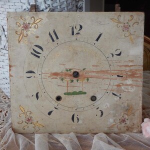 May include: A square, weathered clock face featuring black Arabic numerals from one to twelve arranged in a circle. The off-white surface displays faded floral corner motifs in gold and pink, with a central painted landscape scene and three holes.