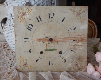 May include: A square, weathered clock face featuring black Arabic numerals from one to twelve arranged in a circle. The off-white surface displays faded floral corner motifs in gold and pink, with a central painted landscape scene and three holes.