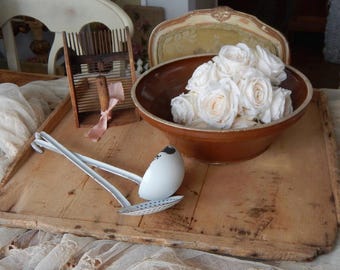 May include: A wooden tray holding a brown ceramic bowl filled with white artificial roses, a wooden wool comb with a pink ribbon, and two white enameled metal ladles, one of which features a perforated bowl for straining liquids.
