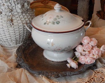 May include: A white enamel tureen with a lid, featuring hand-painted pink rose motifs and green foliage. The round vessel has two side handles, a pedestal base, and a dark red rim. It sits on a dark, ornate metal tray next to a cluster of pink flowers.