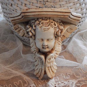 May include: A cream-colored wall shelf bracket featuring a cherub face with detailed wings. The bracket has a distressed finish, with brown accents. The shelf is mounted on a white wicker basket, resting on a white lace fabric.