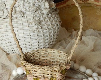 May include: A small, woven basket with a curved handle. The basket is decorated with pink and green floral accents around the base. A larger white basket with floral details is in the background.
