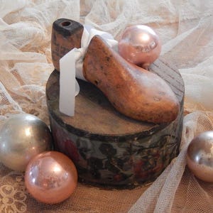 May include: A wooden shoe last and a small wooden block are atop a round, antique box with floral designs. Several glass ornaments in silver and rose gold hues surround the box. A white ribbon is tied around the shoe last.