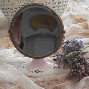 May include: A vintage vanity mirror with a round, reflective surface and a brass frame. The base is pink and white porcelain, decorated with floral patterns. Dried purple flowers rest beside the mirror, on a lace cloth.