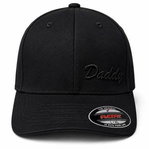 May include: A solid black baseball cap featuring the word Daddy embroidered in a matching black script font on the front panel. The cap includes a curved brim and a circular sticker on the right side that reads The Original Flexfit L/XL.