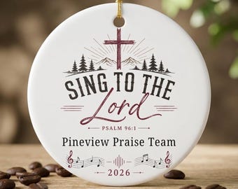 Puede incluir: Un adorno redondo de cerámica blanca con una cruz granate sobre un gráfico de montañas y pinos. El texto dice "SING TO THE LORD", "PSALM 96:1", "Pineview Praise Team" y "2026". Se incluyen notas musicales y un cordón dorado para colgar.