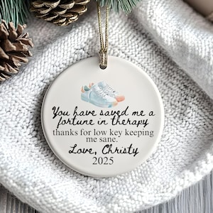 Puede incluir: Adorno navideño redondo de cerámica blanca con una cuerda dorada para colgar. Presenta una imagen de acuarela de zapatillas deportivas y el texto "You have saved me a fortune in therapy...Love, Christy 2025."