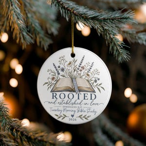 May include: A round white ceramic ornament featuring an illustration of an open book with wildflowers emerging from the pages. The text reads ROOTED and established in love EPHESIANS 3:17 Tuesday Morning Bible Study 2026 with a small heart icon.