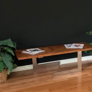 May include: A long, rectangular wooden coffee table with a natural wood finish and a silver metal base. The table is sitting on a hardwood floor and has a few magazines on top.