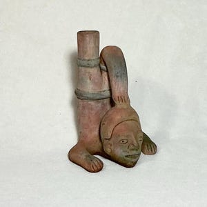 May include: A terracotta sculpture of a figure in a handstand pose. The figure has a cylindrical body with two bands around it, a head with facial features, and small feet and hands. The sculpture is primarily reddish-brown with gray accents.