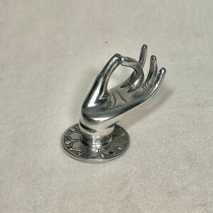 May include: A silver-colored hand sculpture in the mudra position, mounted on a circular base with a floral design. The hand is detailed with lines and curves, and the base has a small hole. The sculpture is likely made of metal.