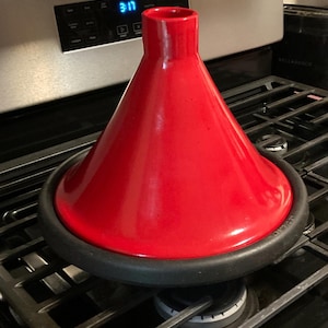 May include: A vibrant red tagine pot with a conical lid and a black base, sitting on a gas stove. The pot's design is traditional, and the bright color makes it a striking kitchen item. The stove's digital display shows the time.