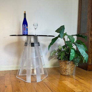 May include: A clear acrylic table with a round top and a unique, geometric base. A bottle of wine and a wine glass are on the table. A potted plant with green leaves is in the background.