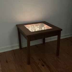 May include: A square wooden side table with a dark brown finish. The tabletop has a recessed glass panel filled with small, warm white fairy lights. The table has four straight legs and is set against a neutral wall.