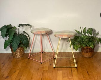 May include: Two metal stools with colorful patterned fabric seats. The stool on the left has a pink metal frame and the stool on the right has a yellow metal frame.