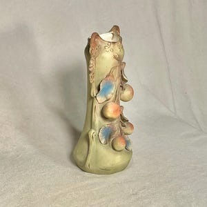 May include: A tall, light green ceramic vase with a narrow neck and flared base. The vase is adorned with sculpted branches, leaves, and fruit in shades of orange, blue, and green. The vase has a height of approximately 8 inches.