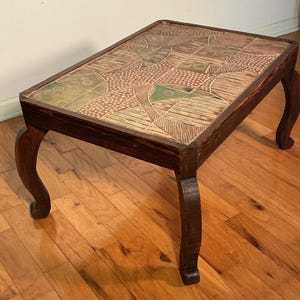 May include: A rectangular wooden coffee table with four curved legs and a dark brown finish. The tabletop features a decorative inlay of ceramic tiles with a geometric pattern in shades of tan, brown, and muted green, showcasing intricate line work.