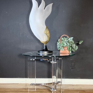 May include: A clear acrylic side table with a glass top and a chrome base. A white and gold abstract sculpture sits on the table, and a small potted plant is next to it.