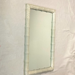 May include: Rectangular mirror with a white, textured frame designed to resemble bamboo. The frame has a distressed, crackled finish. The mirror's surface is clear, reflecting the surrounding environment. The mirror is a decorative item.