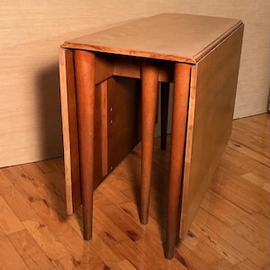 May include: A light brown, mid-century modern drop-leaf table. The table has a rectangular top and four tapered legs. The table is made of wood and has a natural finish. The table is partially folded, showing the drop-leaf design.