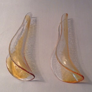 May include: Two amber glass wall sconces with a wavy, organic design. The sconces are made of clear glass with a textured surface and have a warm amber hue.