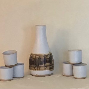 May include: A set of six white ceramic cups and a white ceramic bottle with a brown design. The cups are stacked in two groups of three. The bottle is taller than the cups and has a wide base.