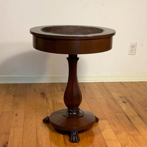 May include: A dark brown wooden side table with a round top and a glass insert. The table has a single pedestal base with a fluted design and four paw feet.
