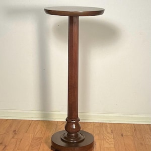 May include: A tall, dark brown wooden pedestal stand featuring a smooth, circular top surface supported by a slender, straight central column. The base is a thick, circular disc with a decorative molded edge, resting on small, rounded feet.