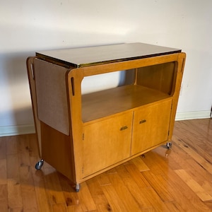 May include: A vintage wooden bar cart with a light wood finish. The cart features a rectangular top, a lower shelf, and a cabinet with two doors. It has a gold-colored trim and sits on small caster wheels.