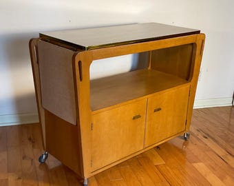 May include: A vintage wooden bar cart with a light wood finish. The cart features a rectangular top, a lower shelf, and a cabinet with two doors. It has a gold-colored trim and sits on small caster wheels.