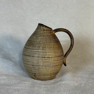 May include: A brown and cream ceramic pitcher with a textured, striped design. The pitcher has a rounded body and a curved handle.