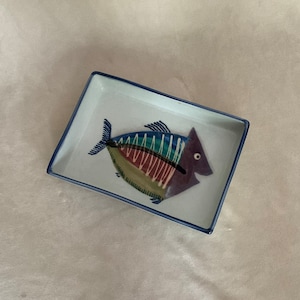May include: A white rectangular ceramic dish with a blue rim features a colorful fish design. The fish is painted in shades of blue, green, purple, and yellow with white accents.