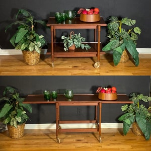 May include: A wooden serving cart with two tiers and wheels. The cart is brown and has a glass top. There are green glasses and a bowl of red fruit on the top tier. The bottom tier has a bowl of red fruit and a green plant.