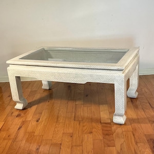 May include: A rectangular coffee table with a glass top and a textured, off-white base. The table has four curved legs and a decorative border around the glass. The table is set on a hardwood floor.