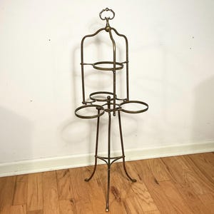 May include: An antique brass plant stand with a decorative top and multiple circular holders. The stand has a tall, slender design with three legs and a series of rings for holding plants or other items. The stand is made of brass and has a vintage aesthetic.