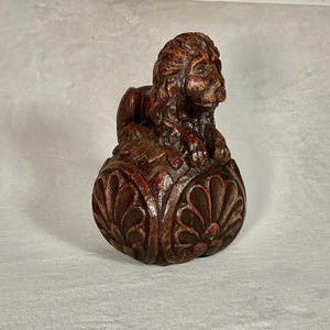 May include: A dark brown wooden carving of a lion resting atop a rounded base. The lion is depicted with a detailed mane and facial features. The base features a carved, repeating petal or leaf pattern that encircles the lower portion of the object.