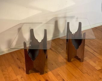 May include: Two matching side tables with dark wood bases and clear glass tops. The bases have a unique, curved design. The tables are placed on a hardwood floor, and the background is a neutral-colored wall.