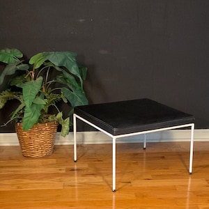 May include: A white metal square ottoman with a black leather top. The ottoman is sitting on a wooden floor.