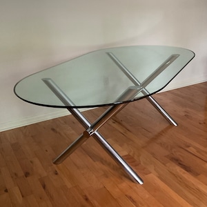 May include: A glass oval-shaped table with a chrome X-shaped base. The table is on a wooden floor.