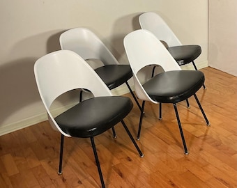 May include: Four mid-century modern chairs. Each chair has a white, curved back with a circular cutout and a black, padded seat. The chairs have black metal legs with silver-colored feet. The chairs are arranged on a hardwood floor.