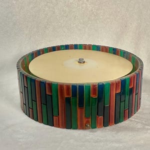 May include: A round flush mount ceiling light fixture featuring a central circular beige panel with a small metal knob. The outer perimeter is composed of vertical, rectangular glass tiles in alternating shades of red, green, and blue.
