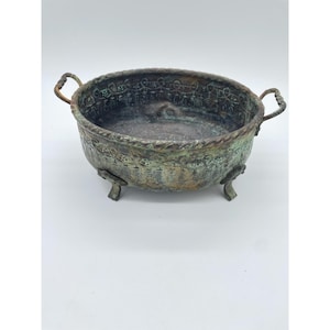May include: An oval metal bowl with a weathered, oxidized green and bronze patina. The bowl features two small side handles, a rope-textured rim, and three short, curved feet. The exterior surface is decorated with an embossed repeating pattern.