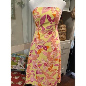 May include: A strapless, knee-length dress with a vibrant floral pattern. The dress features a yellow background adorned with pink, orange, and fuchsia flowers and leaves. The dress is displayed on a mannequin.