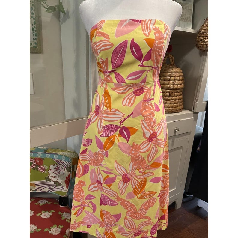 May include: A strapless, knee-length dress with a vibrant floral pattern. The dress features a yellow background adorned with pink, orange, and fuchsia flowers and leaves. The dress is displayed on a mannequin.