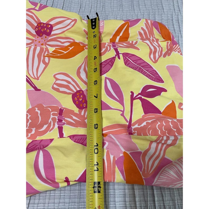 May include: A yellow fabric with a floral pattern in pink and orange. A measuring tape shows the fabric is approximately 12 inches long. The fabric has a repeating pattern of large flowers and leaves.