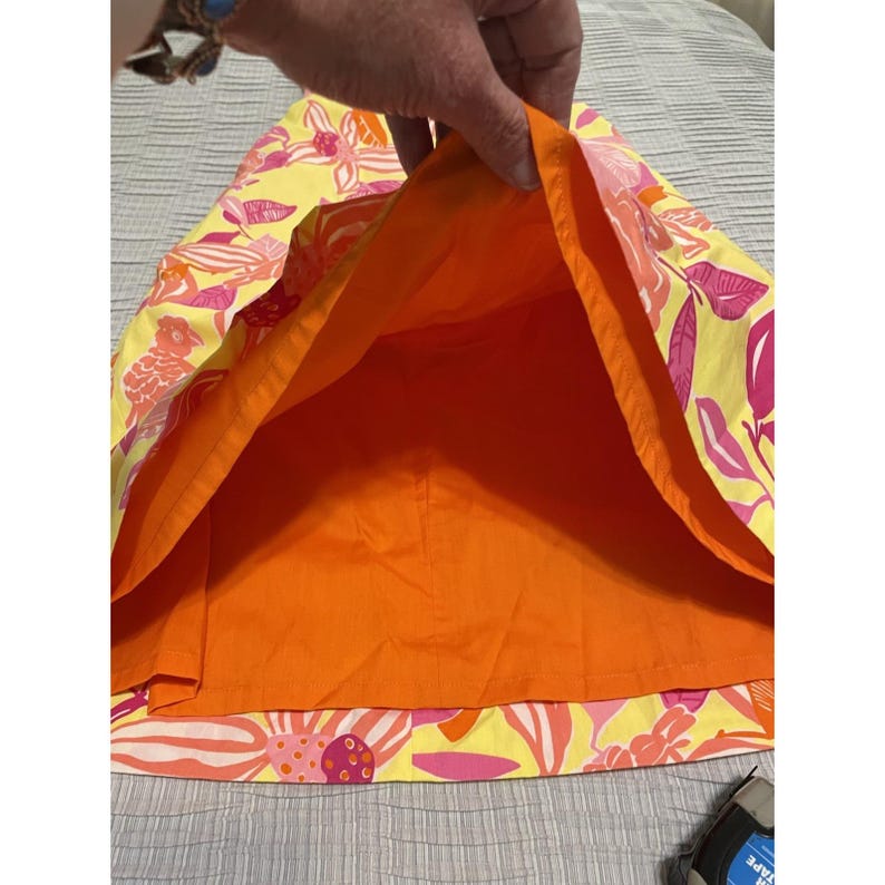 May include: A yellow skirt with a vibrant floral pattern in pink and orange. The skirt's interior is a solid bright orange. The skirt is held up, revealing the interior lining and the colorful exterior design.