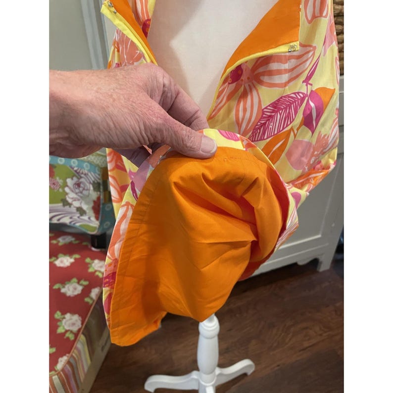 May include: A sleeveless garment with a vibrant floral pattern in yellow, orange, and pink. The inside lining is a solid bright orange. The garment is displayed on a white dress form.