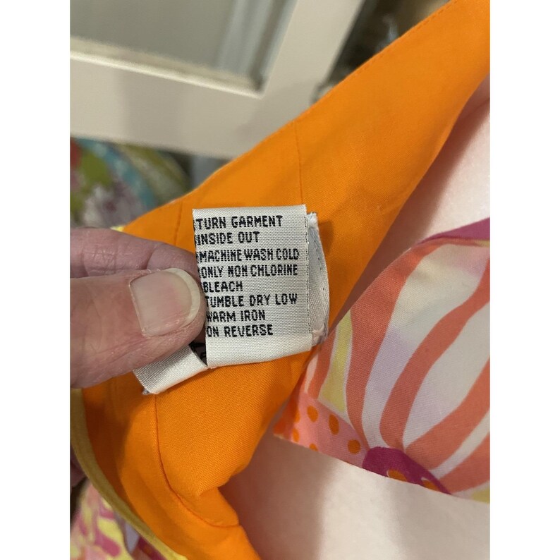 May include: Close-up of a garment tag with care instructions. The tag is white with black text, including "TURN GARMENT INSIDE OUT" and "MACHINE WASH COLD." The garment has an orange lining and a colorful floral pattern.